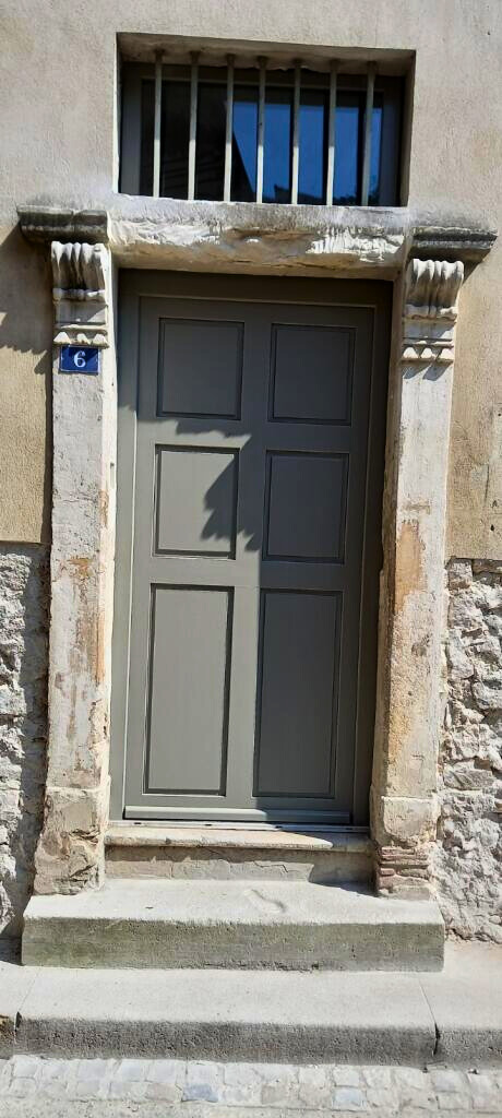 Une porte en bois gris avec des détails architecturaux, encadrée par des pierres anciennes, dans une rue pavée.