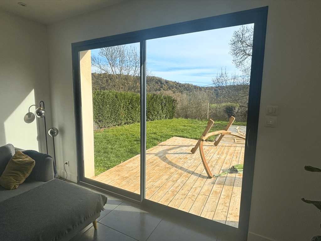 Vue intérieure d'une maison avec une porte-fenêtre donnant sur une terrasse en bois et un jardin vert, avec des collines en arrière-plan.