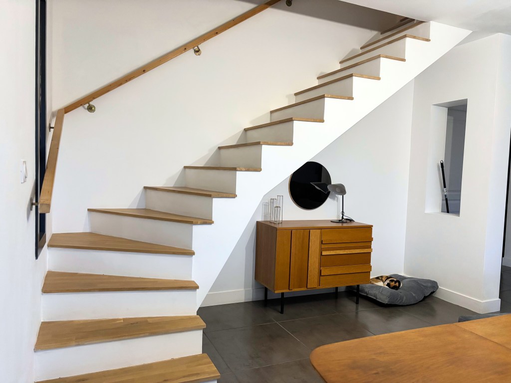 Escalier en bois menant à un étage supérieur, avec un mur blanc et un meuble en bois au bas de l'escalier.