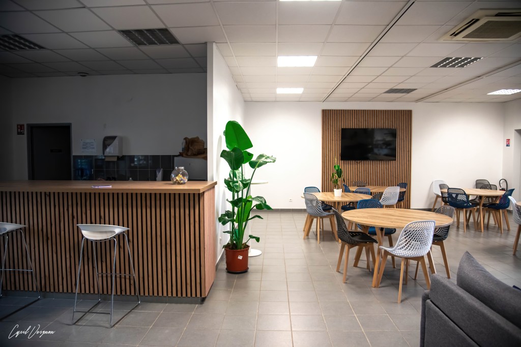Une salle commune moderne avec un bar en bois, des chaises blanches, des tables en bois et des plantes d'intérieur. Un écran est accroché au mur.
