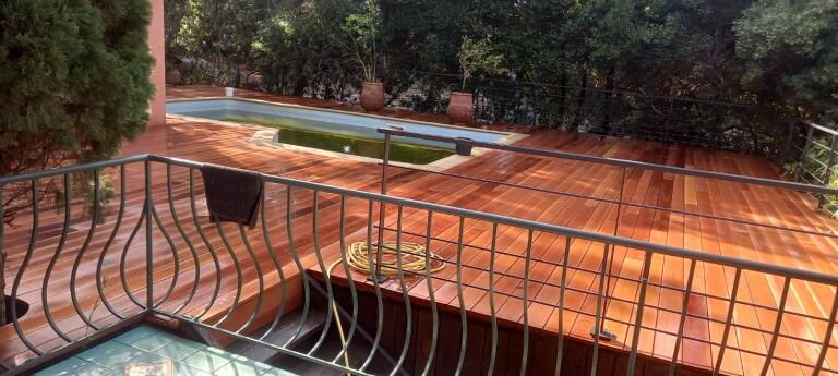 Vue d'un balcon avec une rampe en métal, surplombant une terrasse en bois et une petite piscine, entourée de verdure.
